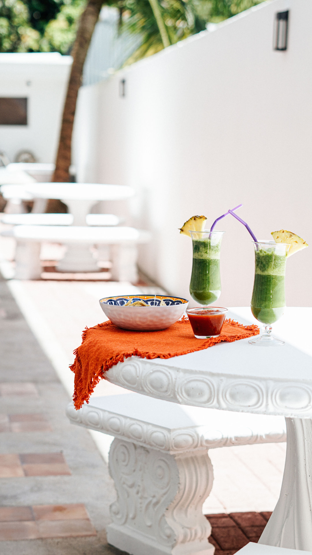 Two green smoothies with pineapple and a bowl of chips on a white poolside table